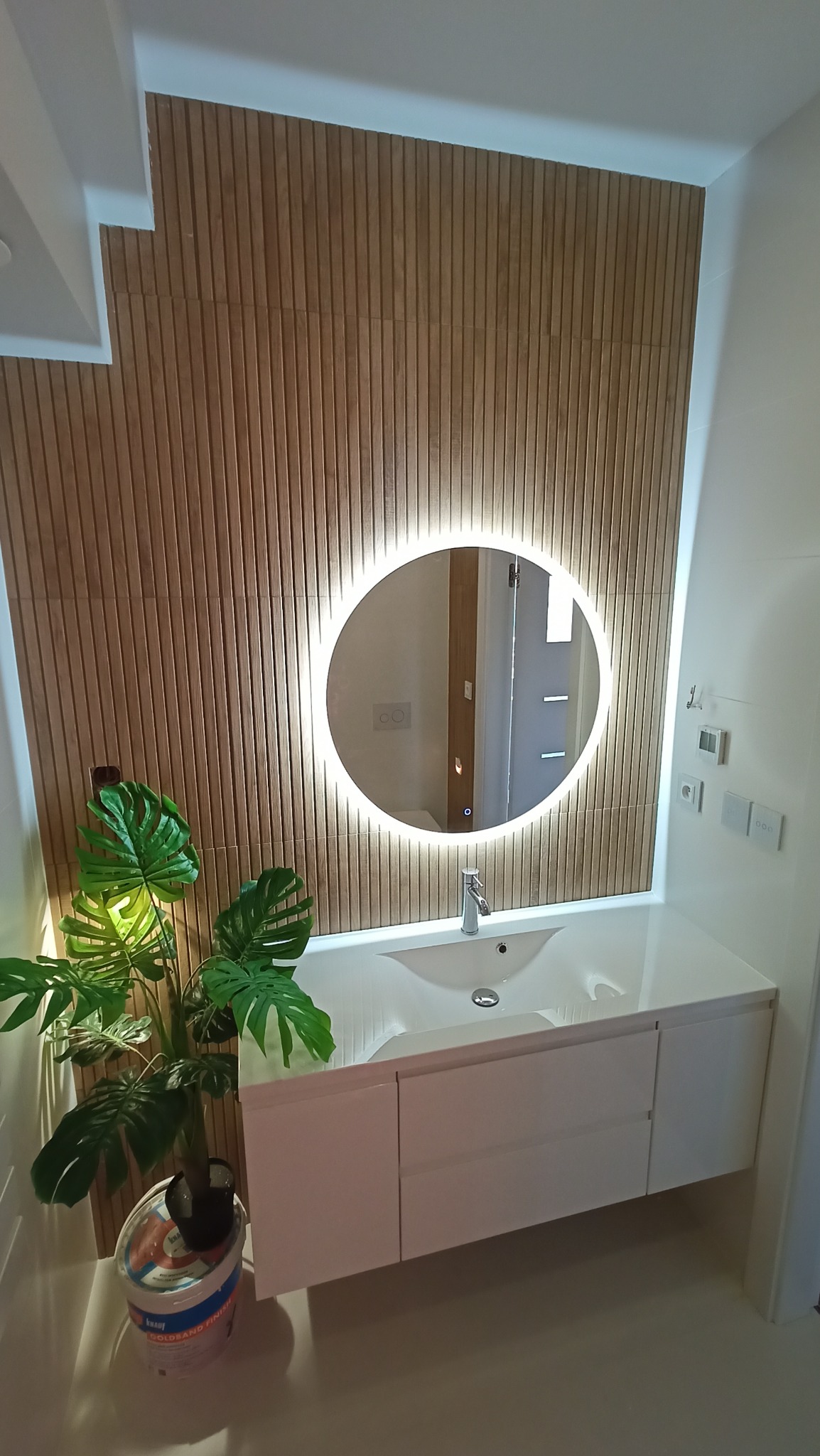 Nowoczesna łazienka z drewnianą ścianą, okrągłym lustrem z podświetleniem LED, białą szafką pod umywalką i rośliną doniczkową obok wiadra z masą szpachlową.