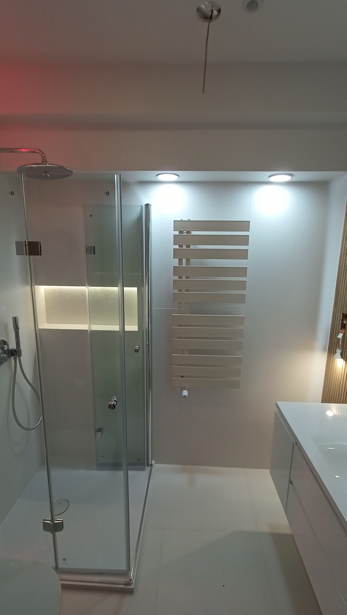 Nowoczesna łazienka w jasnych barwach z kabiną prysznicową ze szklanymi drzwiami, grzejnikiem drabinkowym, umywalką z szafką i wnęką dekoracyjną w ścianie.