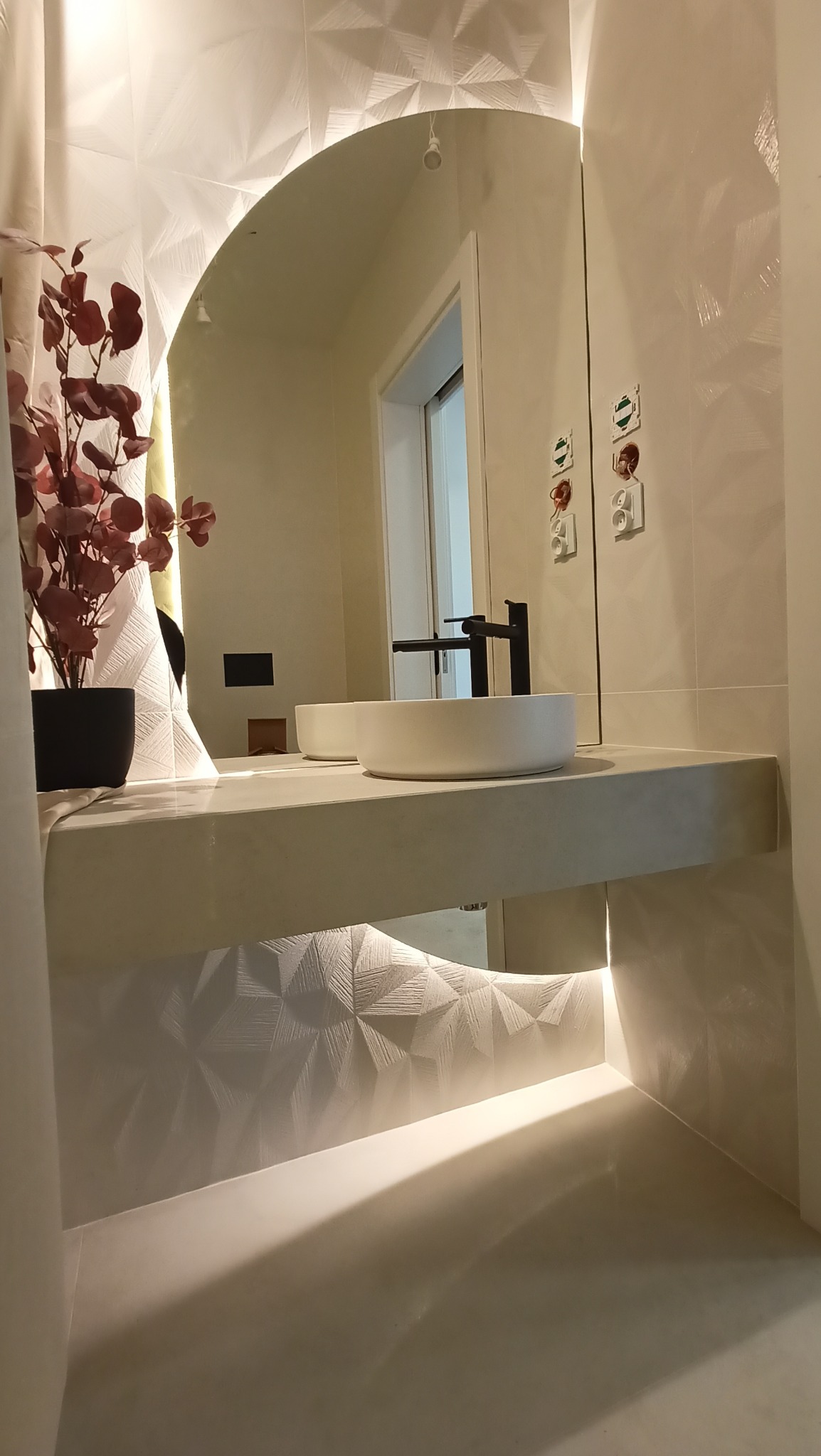 Nowoczesna łazienka z okrągłym lustrem, białą umywalką nablatową i czarną armaturą, podświetlany blat i strukturalna ściana z geometrycznym wzorem.