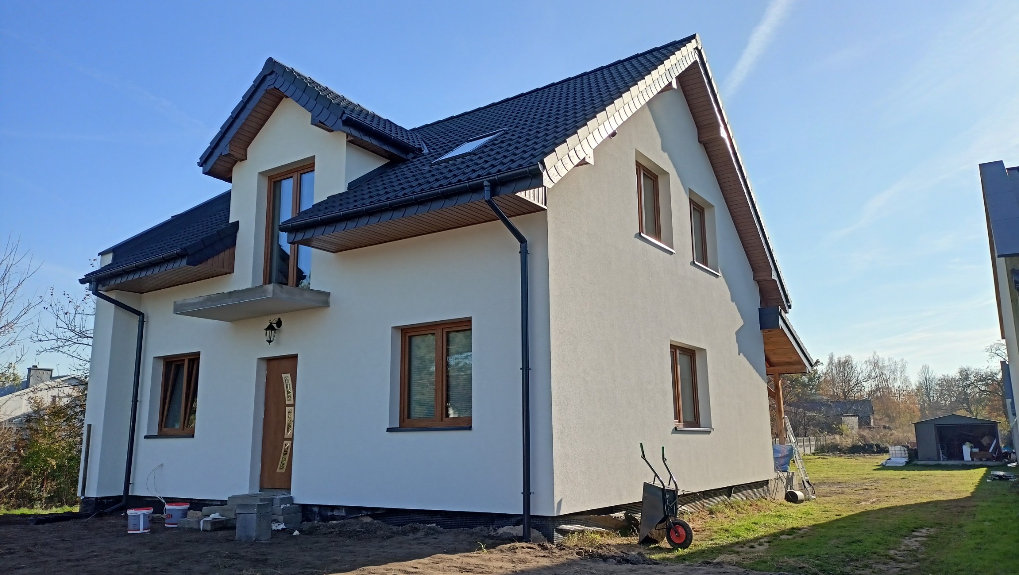Dwupiętrowy dom po ociepleniu elewacji w słoneczny dzień. Widoczne okna z drewnianymi ramami, czarny dach i rynny. Na trawniku stoi taczka i wiaderka z farbą.