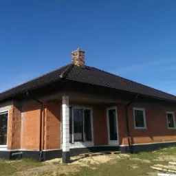Dom jednorodzinny w trakcie budowy: ściany z czerwonej cegły, ciemny dach, białe ramy okienne, widoczny komin z cegły. Błękitne niebo w tle.