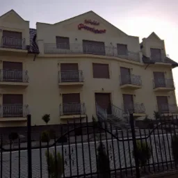 Trzypiętrowy budynek z balkonami i zamkniętymi okiennicami, widoczny zza ozdobnego, kutego ogrodzenia. Nad wejściem widnieje podświetlony szyld z nazwą budynku.