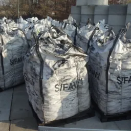 S/FAVI: Szare worki typu big bag z ekogroszkiem na paletach, ustawione na zewnątrz. W tle betonowe prefabrykaty i drzewa bez liści.