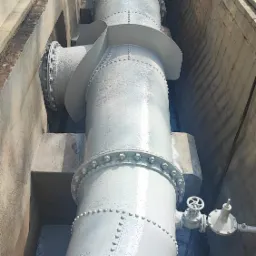 Długi, pomalowany na szaro rurociąg przemysłowy z licznymi łączeniami śrubowymi i zaworami, umieszczony w betonowym kanale.