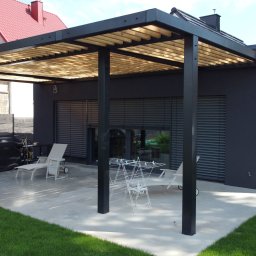 TEKTRUM Sp. z o.o. Sp.k. - Nowoczesna pergola z drewnianym zadaszeniem i czarną konstrukcją na betonowym tarasie przylegającym do ciemnoszarego domu z roletami zewnętrznymi.