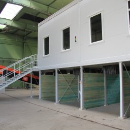 TEKTRUM Sp. z o.o. Sp.k. - Wnętrze hali z podwyższonym, modułowym biurem o białej fasadzie z oknami, wspartym na słupach. Widoczne schody metalowe i fragment transportera taśmowego.