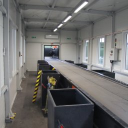 TEKTRUM Sp. z o.o. Sp.k. - Wnętrze hali z linią produkcyjną: szara taśma transportowa, czarne pojemniki, białe ściany i sufit z oświetleniem. Widoczne okna i elementy konstrukcyjne.