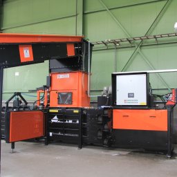 TEKTRUM Sp. z o.o. Sp.k. - Pomarańczowo-czarna prasa hydrauliczna do odpadów w hali produkcyjnej z zielonymi ścianami. Widoczny panel sterowania i osoby w tle.