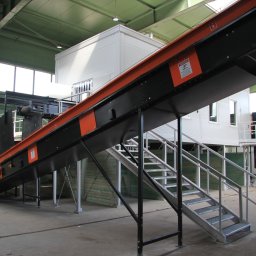 TEKTRUM Sp. z o.o. Sp.k. - Wnętrze hali z konstrukcją stalową: pomarańczowo-czarny podajnik taśmowy na metalowych podporach i schodach. W tle biały budynek kontenerowy i okna.