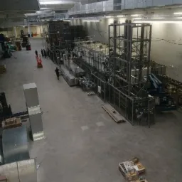 Wnętrze hali produkcyjnej z zainstalowaną linią technologiczną do obróbki, widoczne elementy konstrukcji stalowej, system wentylacji pod sufitem, pracownicy i wózki widłowe w tle.