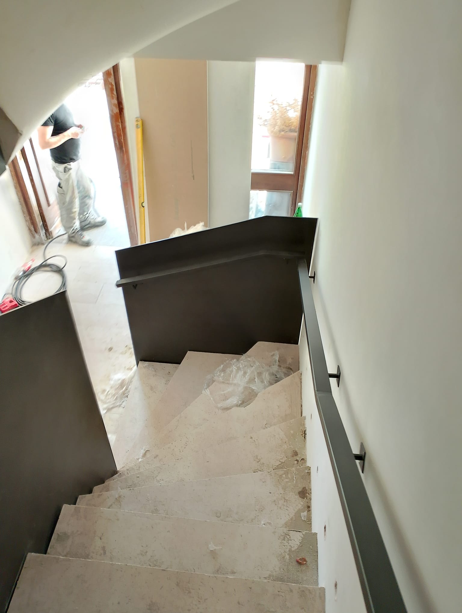 Wewnętrzne schody betonowe w trakcie montażu z szarą metalową balustradą, widoczne narzędzia i pracownik w tle, surowy stan wykończenia.