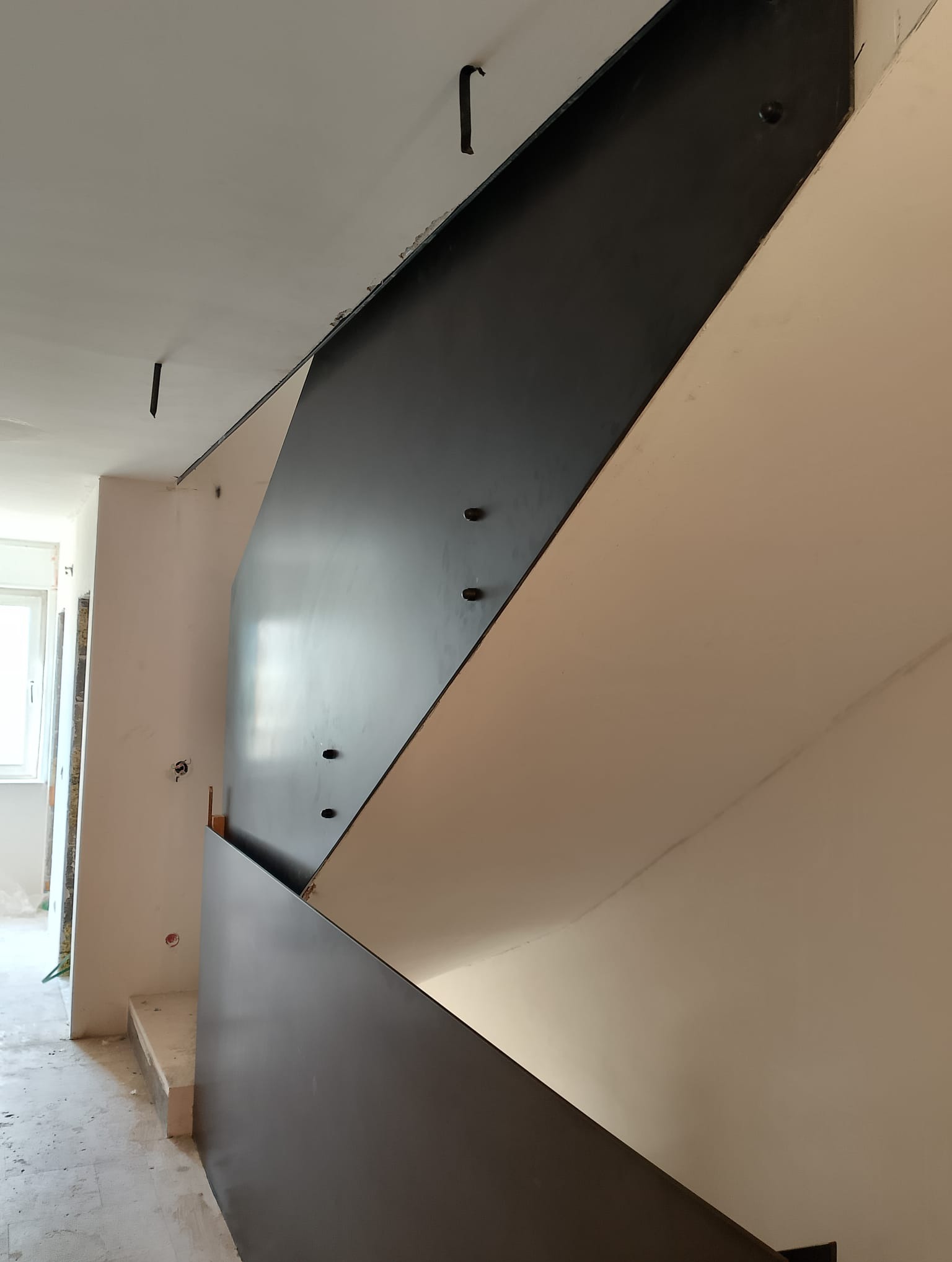 Wnętrze w trakcie wykańczania, widok na metalową konstrukcję schodów z czarnymi panelami bocznymi, zamocowanymi do ściany za pomocą śrub.