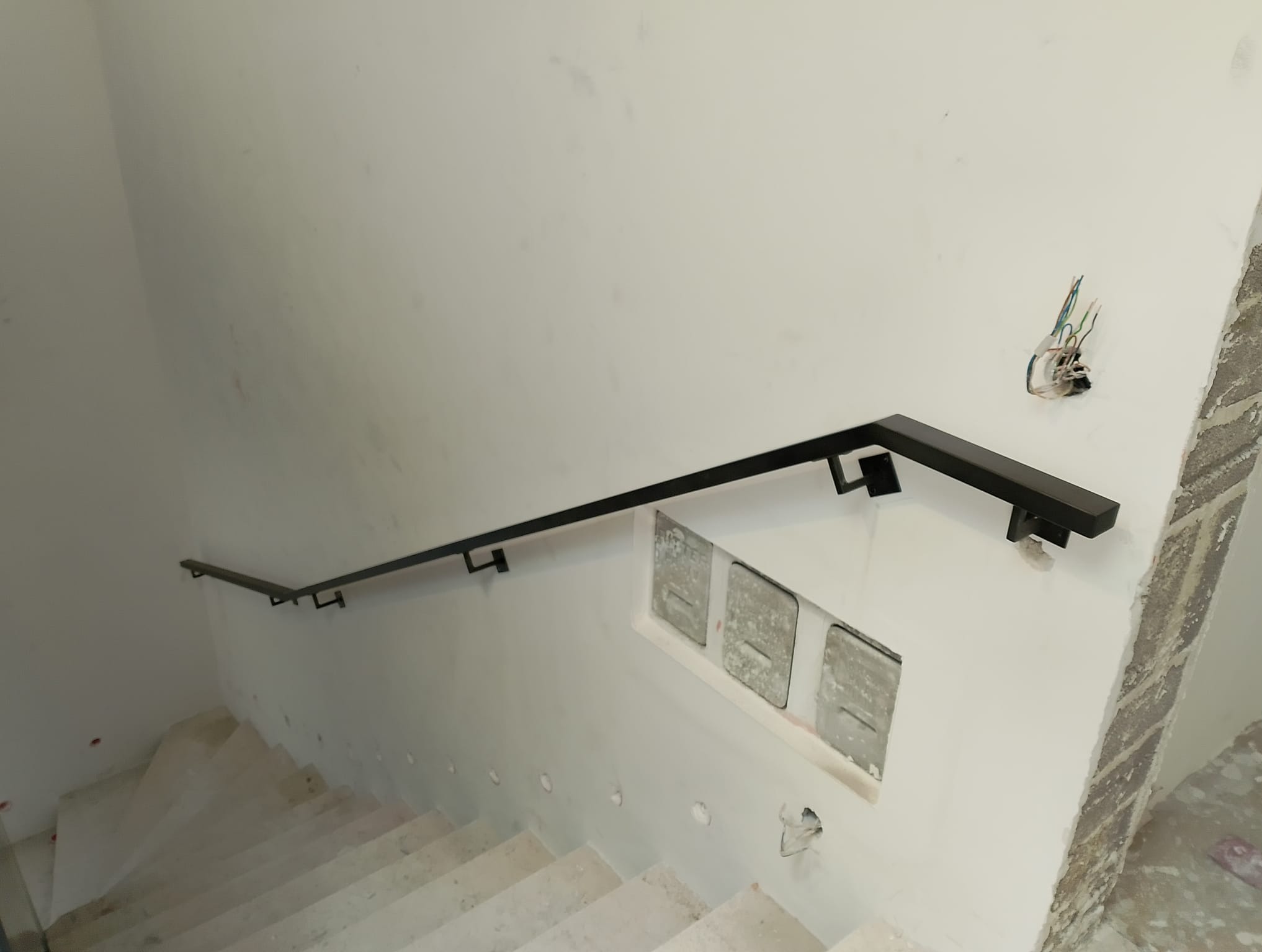 Widok z góry na klatkę schodową w trakcie wykańczania, z zamontowaną, prostą, czarną, metalową poręczą przy ścianie. Widoczne otwory na skrzynki pocztowe i wystające przewody elektryczne.