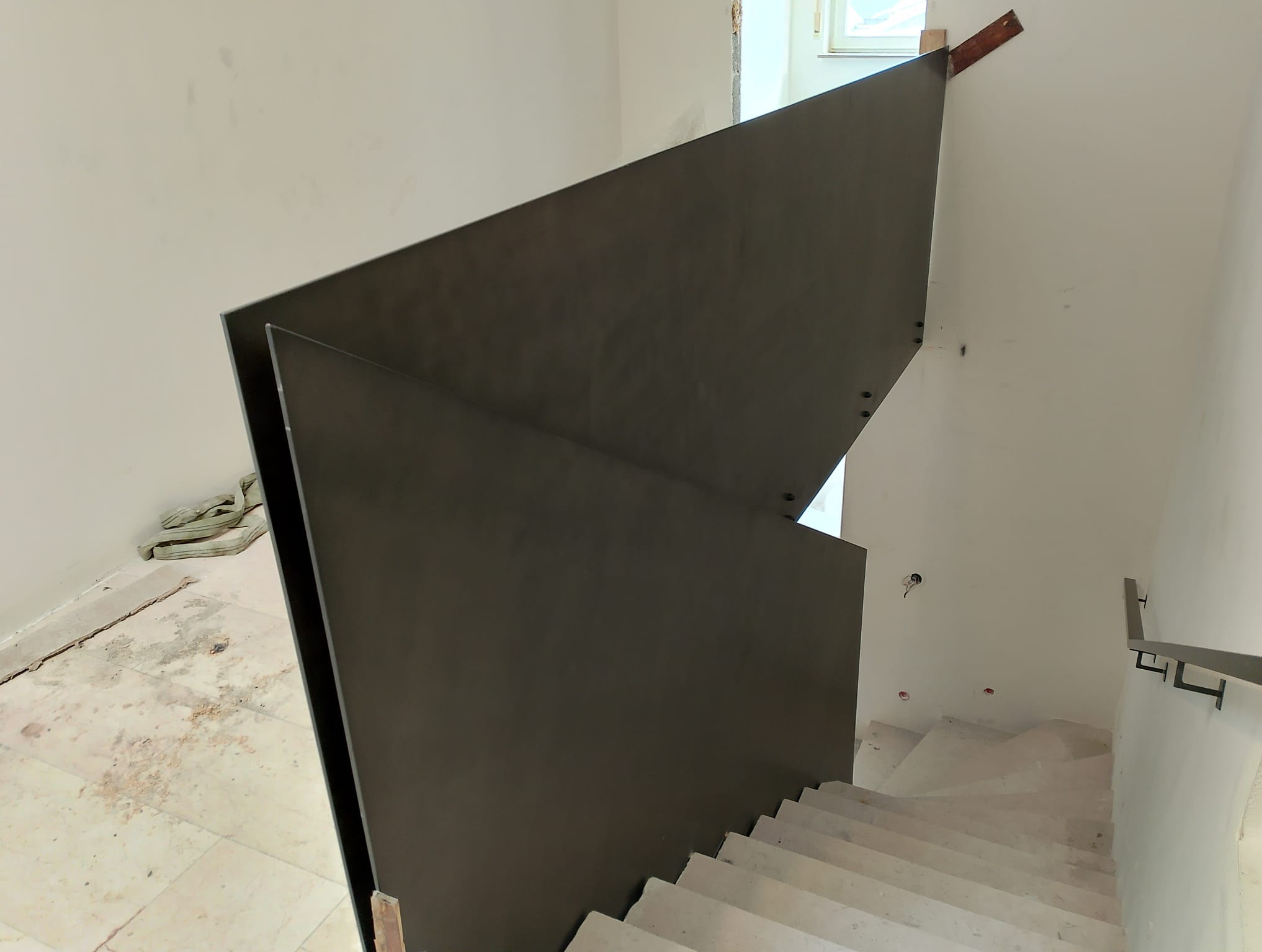 Wnętrze w trakcie wykańczania: surowe betonowe schody z nowoczesną, geometryczną balustradą ze stali w odcieniach szarości, zamocowaną do ściany na drewnianych elementach, widok z góry.