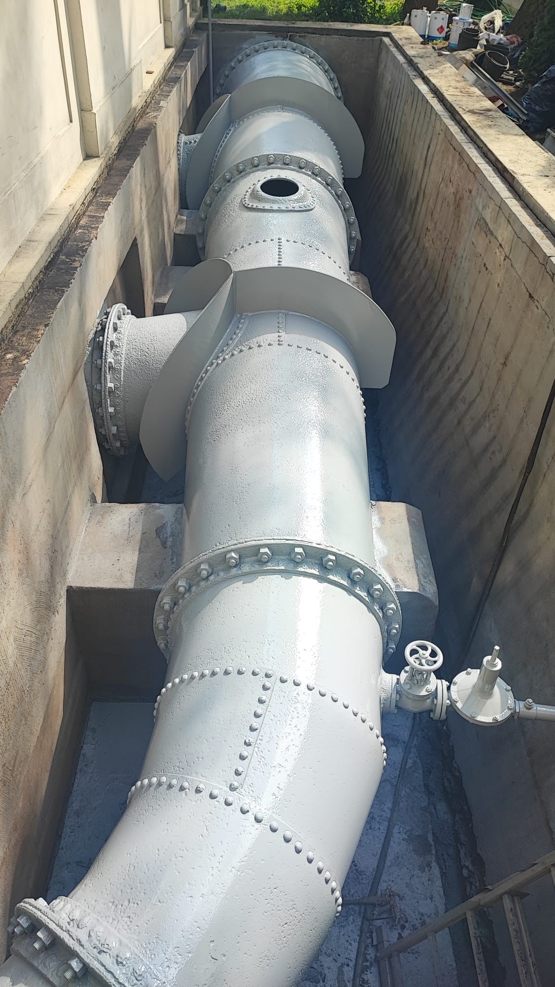 Długi, pomalowany na szaro rurociąg przemysłowy z licznymi łączeniami śrubowymi i zaworami, umieszczony w betonowym kanale.