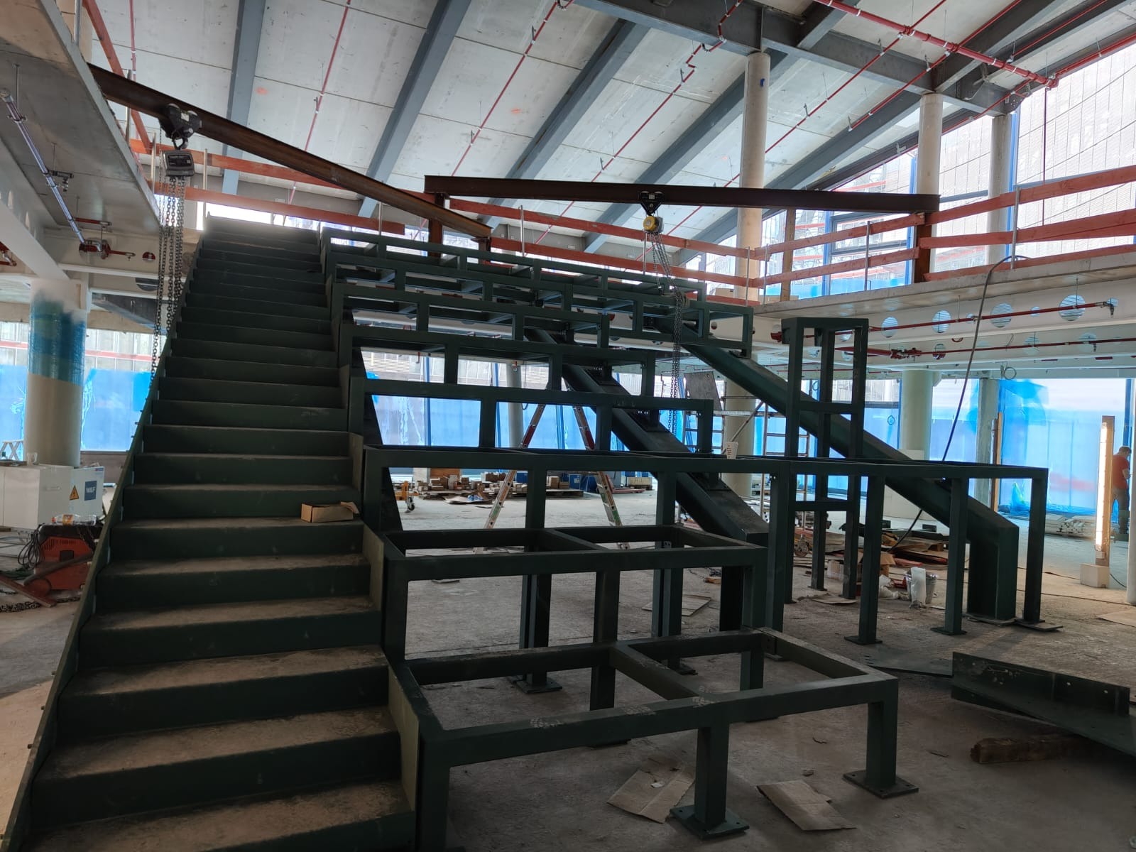 Montaż metalowych schodów w surowym stanie budynku usługowego, widoczne elementy konstrukcyjne i podwieszony łańcuch, w tle okna z niebieską folią ochronną.