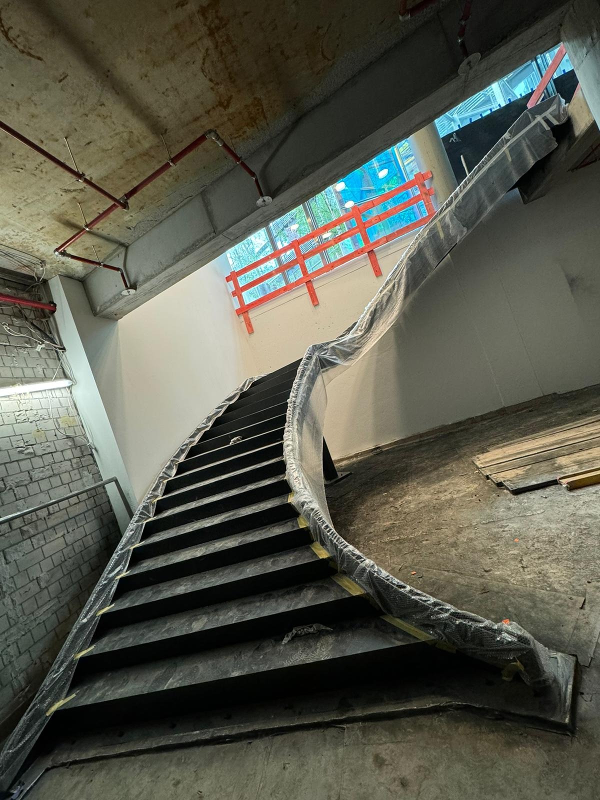 Zakryte folią ochronną, spiralne schody metalowe w surowym stanie, z widocznym oknem z pomarańczową barierką w tle, w trakcie prac wykończeniowych wnętrza.