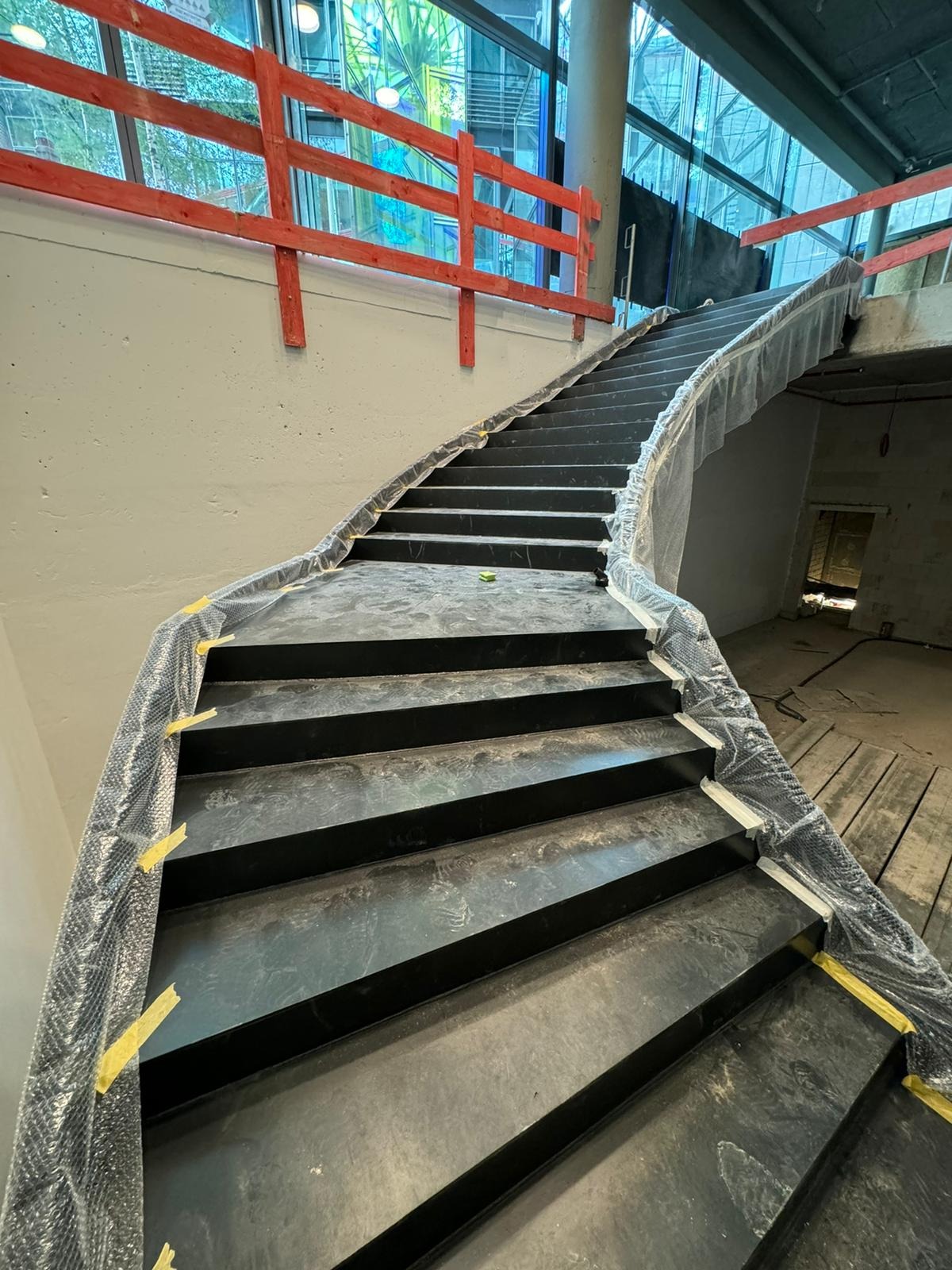 Nowe schody metalowe w trakcie budowy, zabezpieczone folią ochronną i taśmą, widoczne okna z kolorowymi szybami i pomarańczowe barierki.