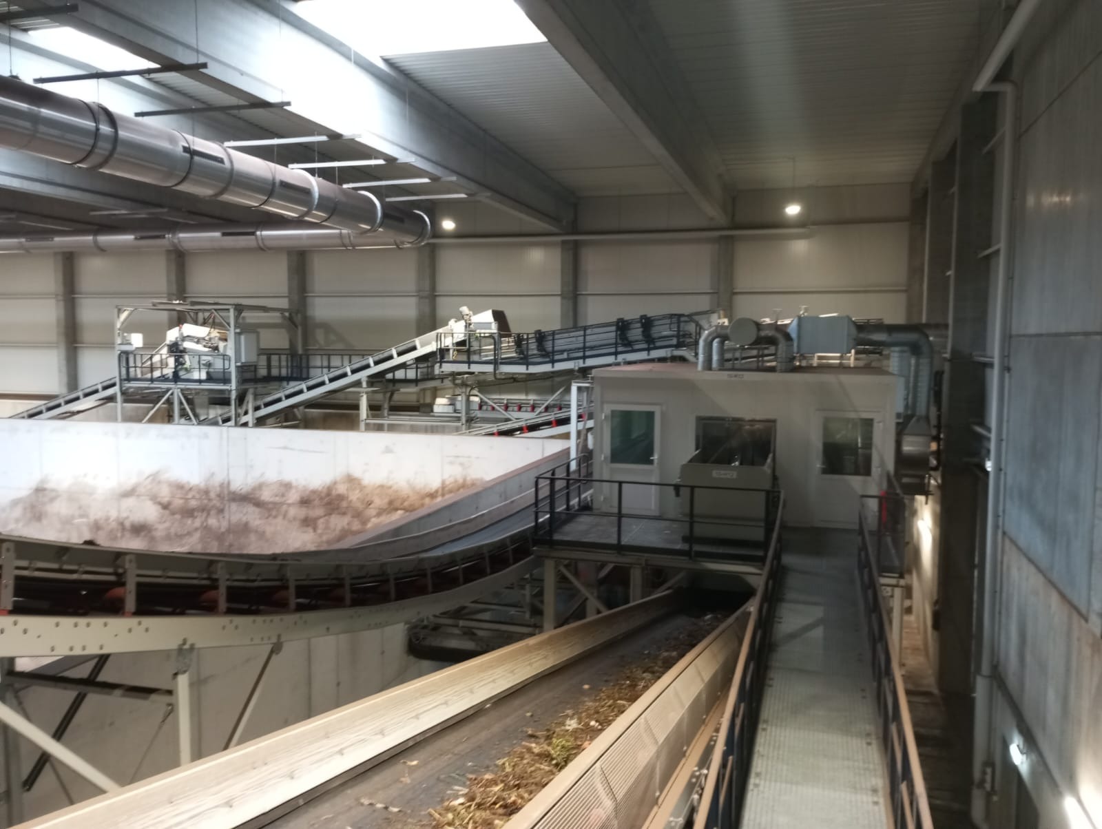 Wnętrze hali przemysłowej z systemem przenośników taśmowych transportujących odpady, widoczna kabina operatorska i stalowa konstrukcja wsporcza.