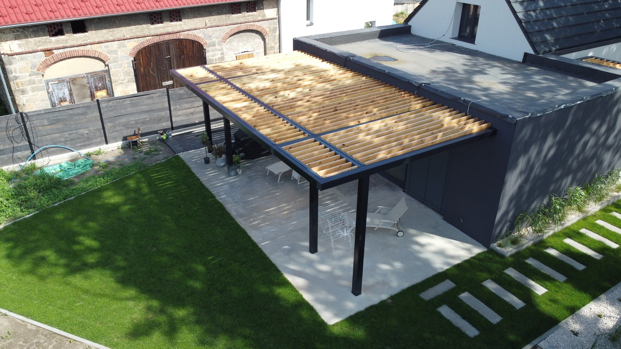 Nowoczesna pergola z drewnianym zadaszeniem i czarną ramą, przylegająca do budynku z płaskim dachem. Widok z góry na zadbany trawnik i ścieżkę z płyt.