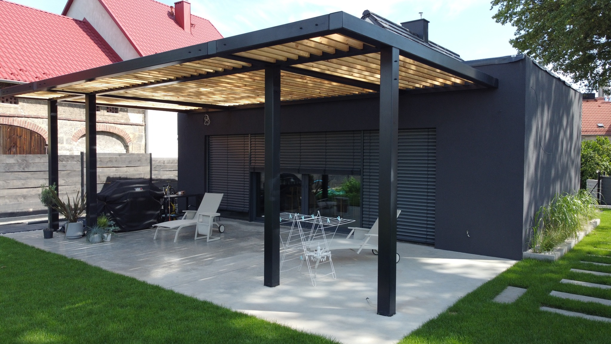 Nowoczesna pergola z drewnianym zadaszeniem i czarną konstrukcją na betonowym tarasie przylegającym do ciemnoszarego domu z roletami zewnętrznymi.