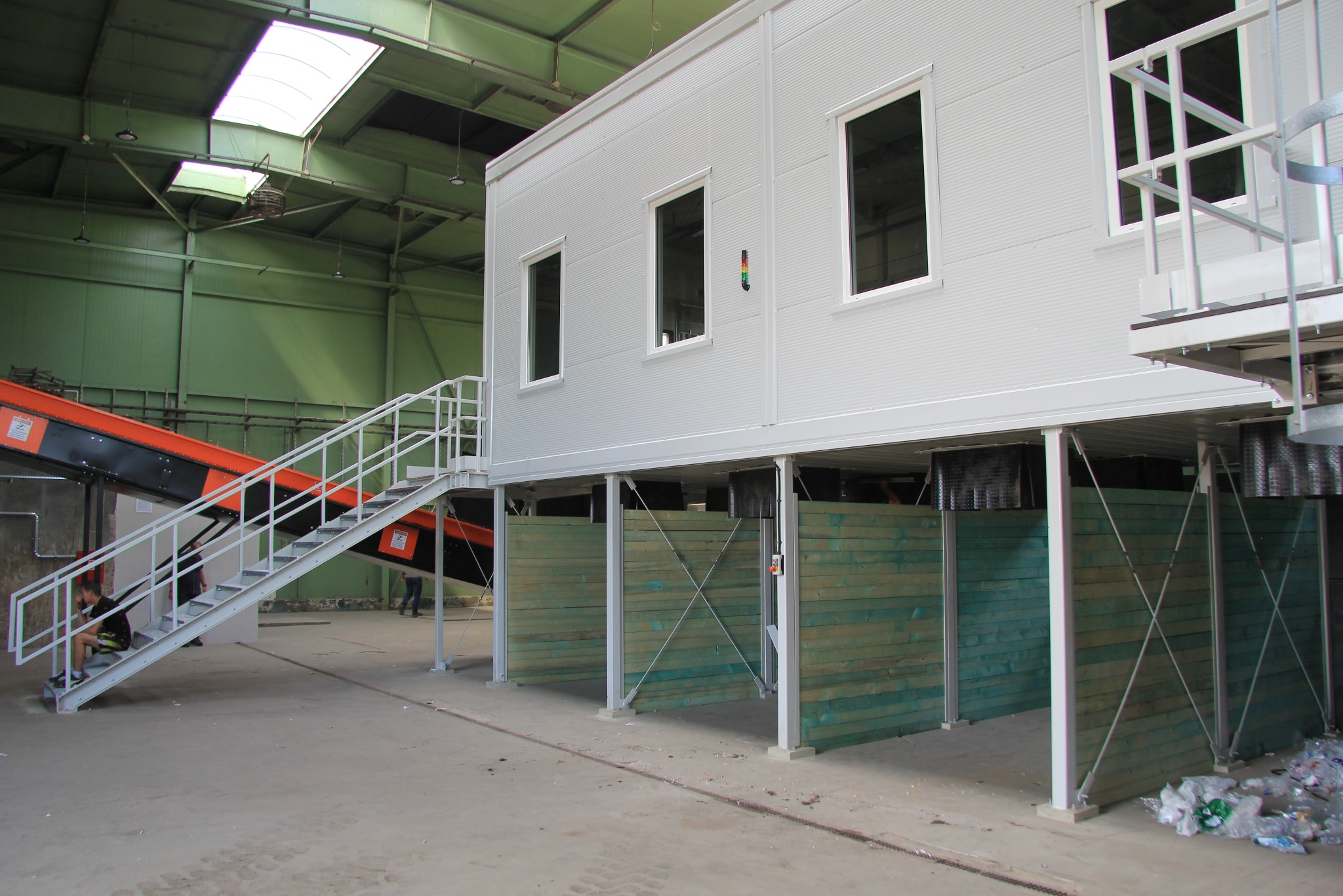 Wnętrze hali z podwyższonym, modułowym biurem o białej fasadzie z oknami, wspartym na słupach. Widoczne schody metalowe i fragment transportera taśmowego.