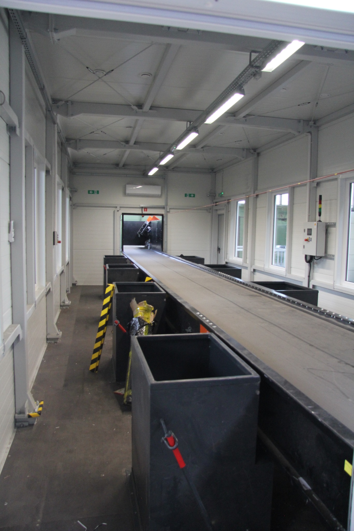 Wnętrze hali z linią produkcyjną: szara taśma transportowa, czarne pojemniki, białe ściany i sufit z oświetleniem. Widoczne okna i elementy konstrukcyjne.