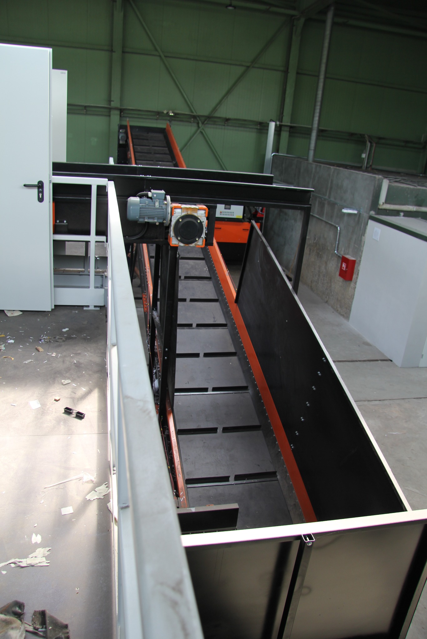 Wnętrze hali z widokiem na metalową konstrukcję przenośnika taśmowego z pomarańczowymi akcentami, oświetloną przez światło wpadające z góry.
