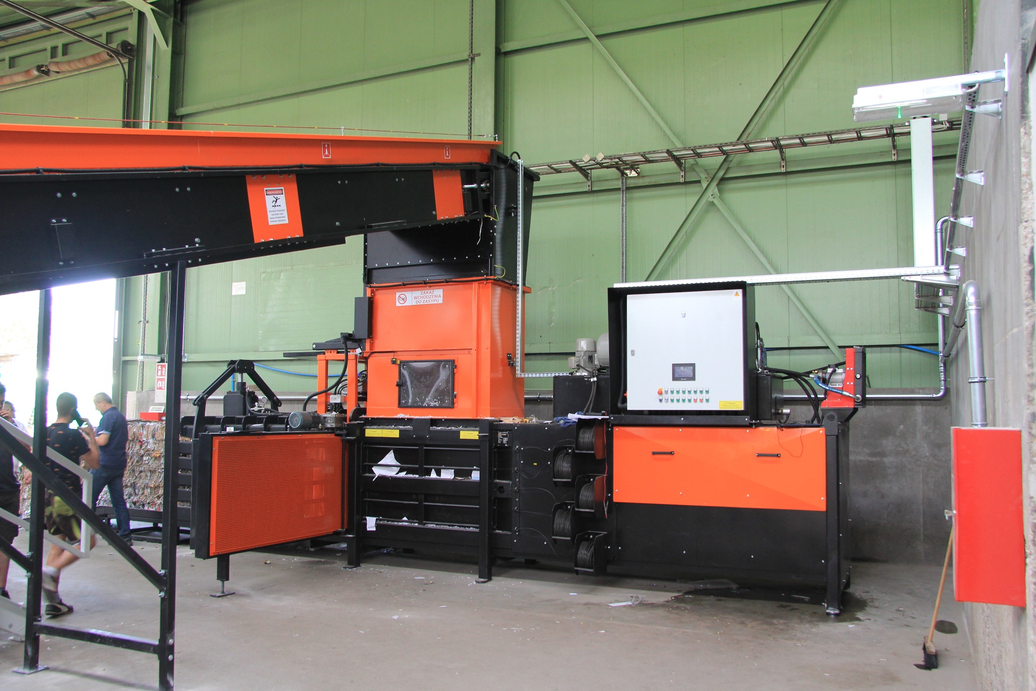 Pomarańczowo-czarna prasa hydrauliczna do odpadów w hali produkcyjnej z zielonymi ścianami. Widoczny panel sterowania i osoby w tle.