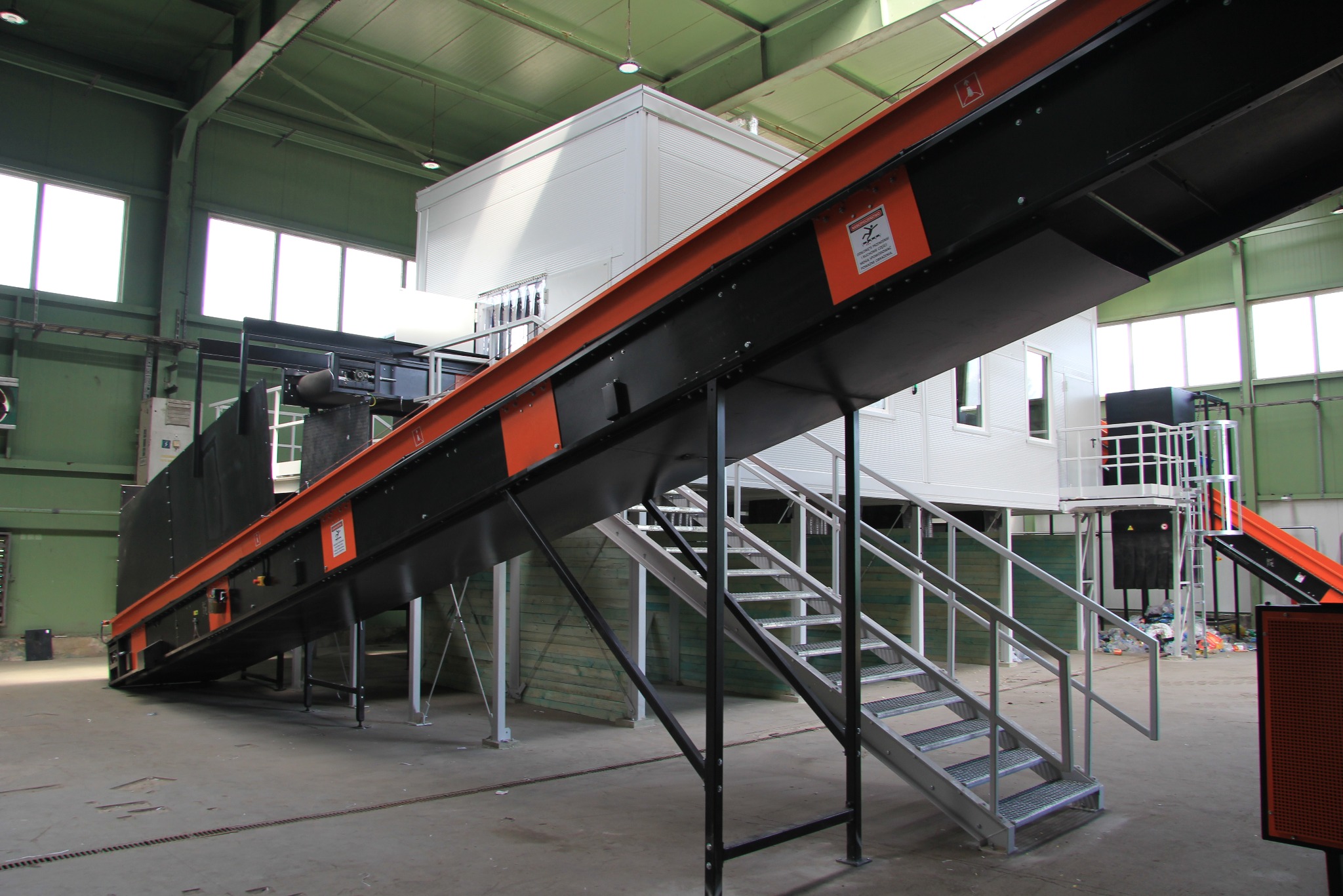 Wnętrze hali z konstrukcją stalową: pomarańczowo-czarny podajnik taśmowy na metalowych podporach i schodach. W tle biały budynek kontenerowy i okna.