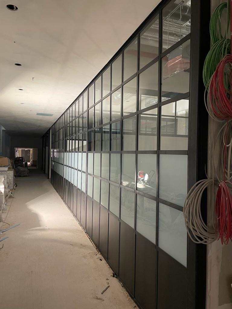 Długi korytarz z przeszklonymi ścianami działowymi w czarnych ramach, widoczne kable i surowy stan wykończenia wnętrza, sugerujący etap prac budowlanych.