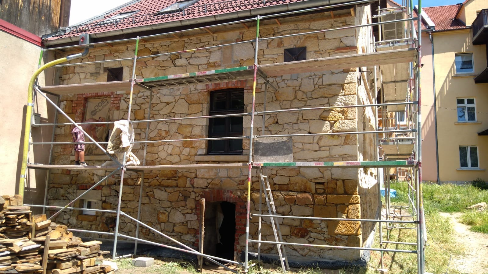 Renowacja kamiennej elewacji budynku z użyciem rusztowań. Widoczne fragmenty nowej stolarki okiennej i przygotowane kamienie do montażu. Prace w toku.