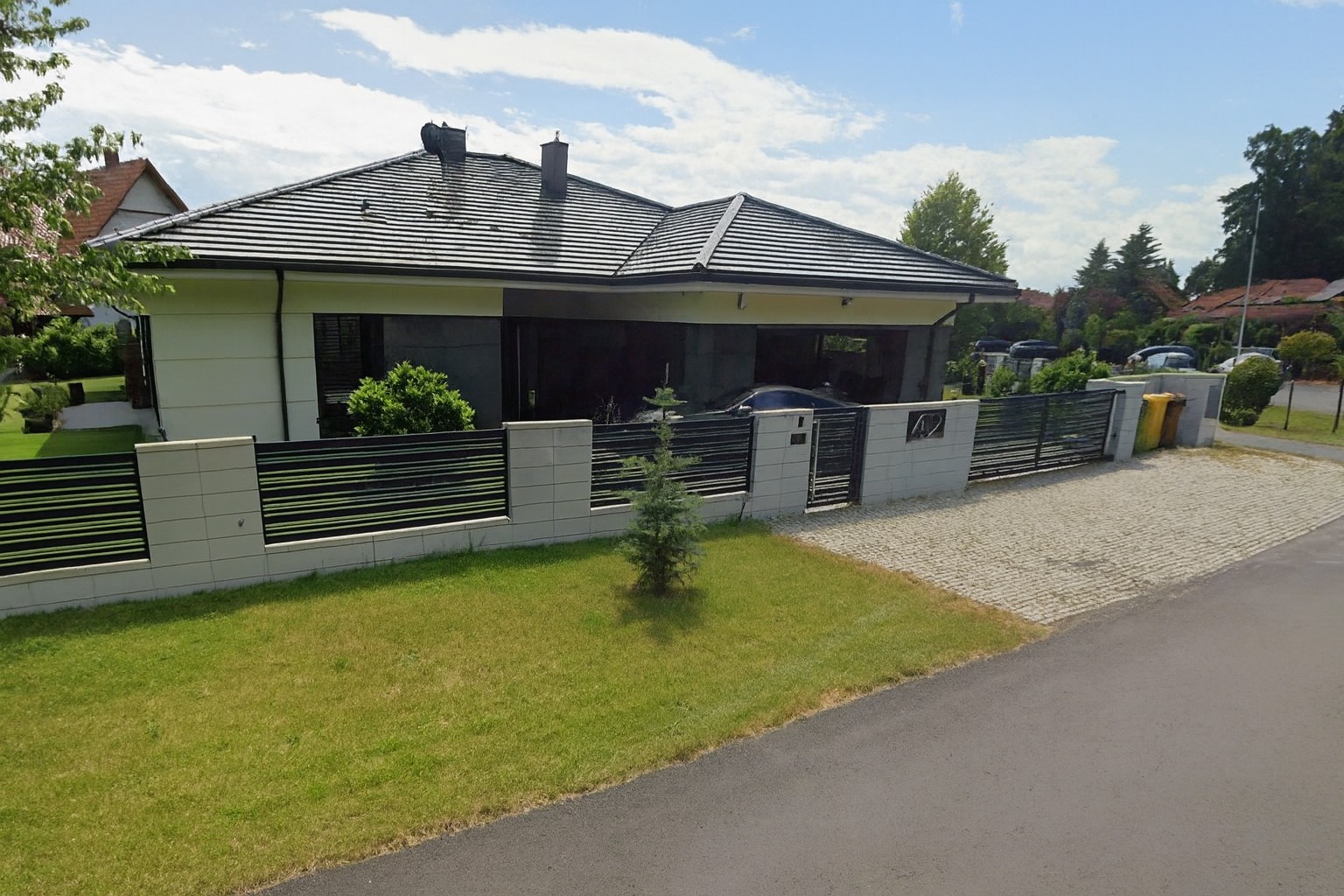 Nowoczesny dom jednorodzinny z płaskim dachem i ogrodzeniem z poziomych paneli, widok z ulicy w słoneczny dzień. Na pierwszym planie trawnik i małe drzewko.