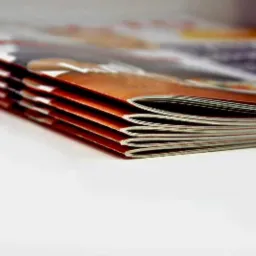 Stos zadrukowanych arkuszy, prawdopodobnie ulotek lub broszur, ułożonych równo na białym tle, widoczne krawędzie z kolorowymi grafikami.