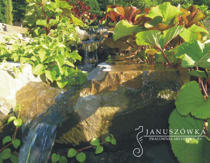 Kaskada wodna w ogrodzie z kamieni i roślin o dużych liściach, w prawym dolnym rogu widoczne logo pracowni architektonicznej.