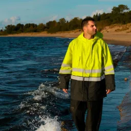 Mężczyzna w jaskrawej, żółto-szarej kurtce roboczej z kapturem, spacerujący brzegiem morza, z bryzgami fal u stóp.
