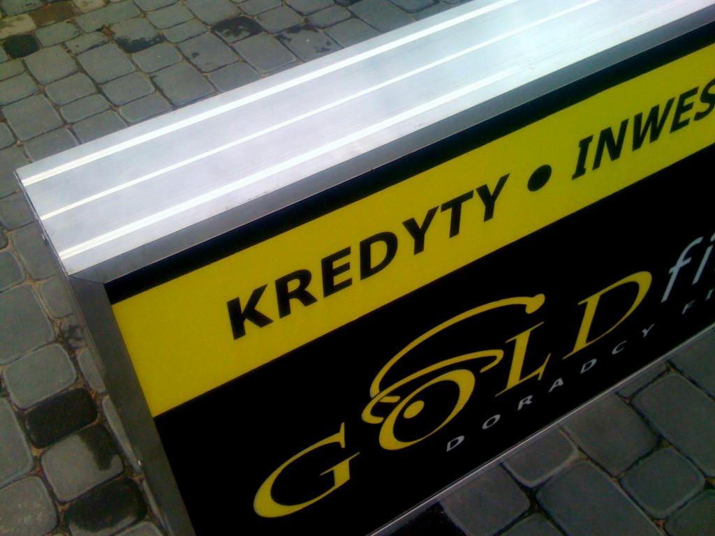 Kaseton reklamowy z żółto-czarnym nadrukiem 'Kredyty Inwestycje Gold fi Doradcy' na tle szarej kostki brukowej, widoczny fragment aluminiowej ramy.