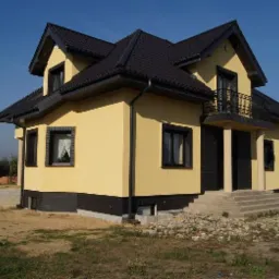 Żółty dom murowany z czarnym dachem i oknami, ocieplony, z balkonem z czarną balustradą, widok z boku na tle błękitnego nieba i zieleni.