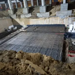 Rozległe zbrojenie fundamentów z widocznymi warstwami siatki stalowej, czarną folią izolacyjną i ścianami wykopu w tle, oświetlone sztucznym światłem budowy.
