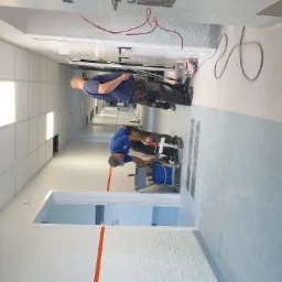 Dw&oacute;ch malarzy w trakcie malowania ścian korytarza w budynku użyteczności publicznej; jeden maluje wałkiem dolną część ściany, drugi maluje g&oacute;rną część ściany za pomocą teleskopowego kija; widoczne...