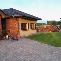 Częściowo ceglany dom z ciemną dachówką, widok z brukowanego podjazdu, z rowerami i dekoracjami ogrodowymi