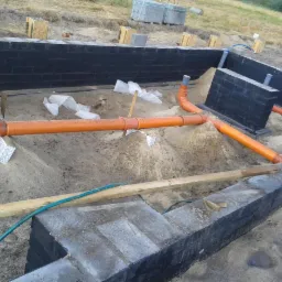 Fundamenty z izolacją i instalacją kanalizacyjną w piasku, widok z góry. Czarna izolacja pionowa na ścianach fundamentowych. Pomarańczowe rury kanalizacyjne w wykopie.
