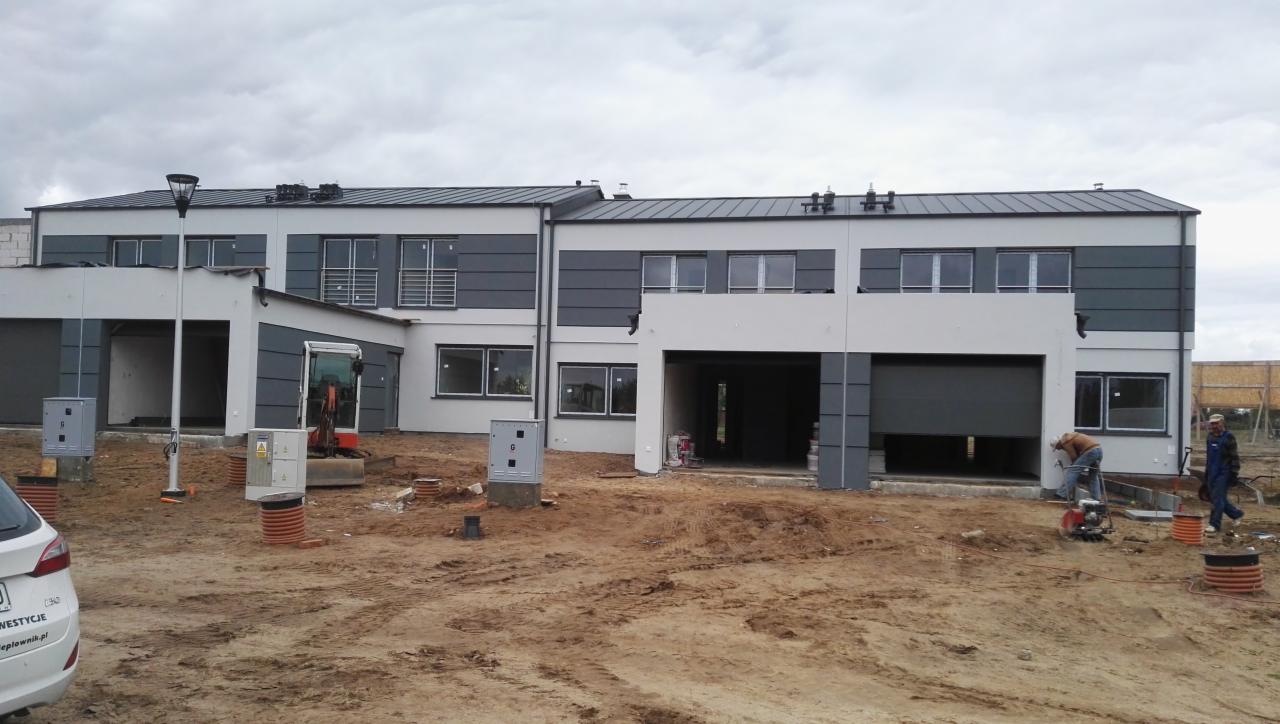 Wznoszone szeregowo domy z garażami, częściowo wykończone elewacje w tonacji szaro-białej. Na pierwszym planie robotnicy i sprzęt budowlany. Częściowo podniesione bramy garażowe.