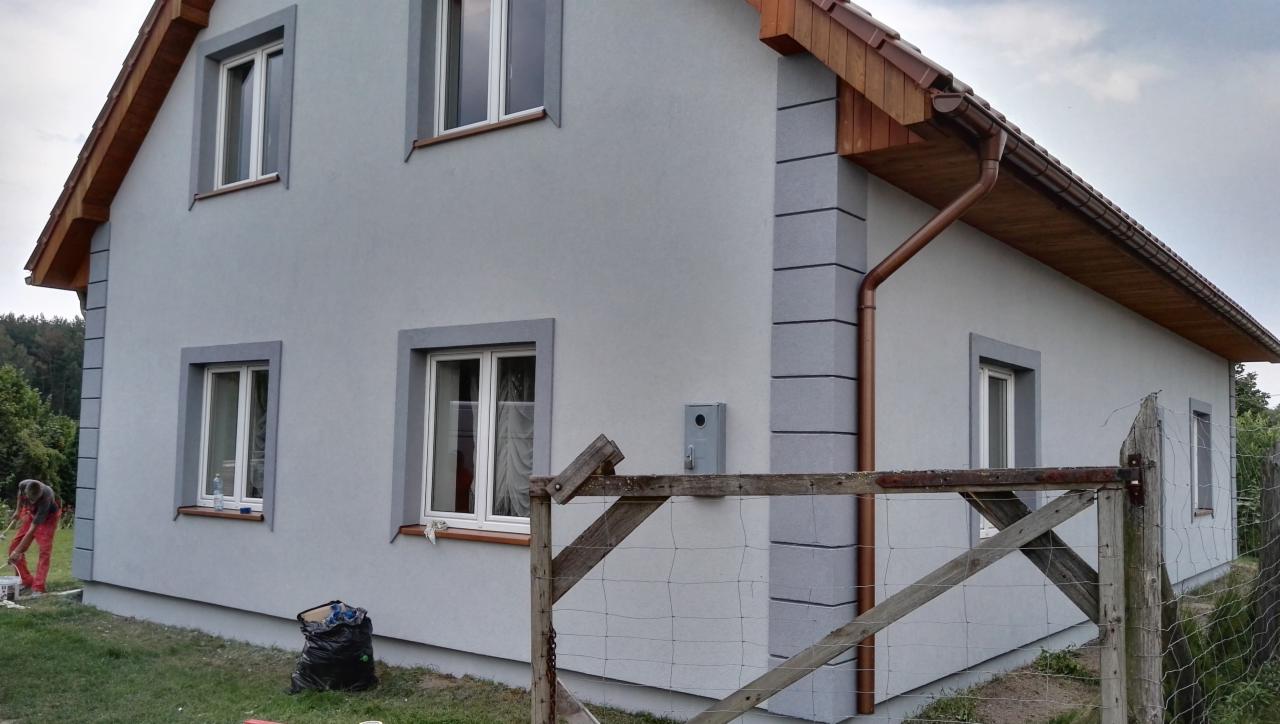 Szary dom jednorodzinny w Szczecinku po remoncie elewacji, widoczne nowe okna z szarymi ramami, drewniana podbitka dachowa i orynnowanie, fragment drewnianego płotu na pierwszym planie.