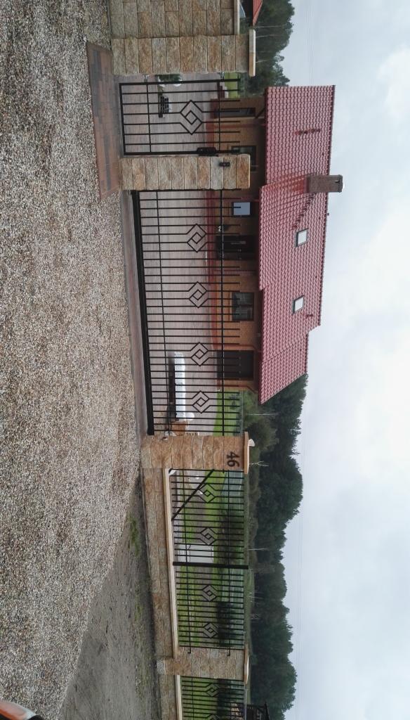 Nowoczesny dom murowany z czerwoną dachówką, widoczny zza czarnej, metalowej bramy z kamiennymi słupkami, na których widnieje numer posesji '46'.