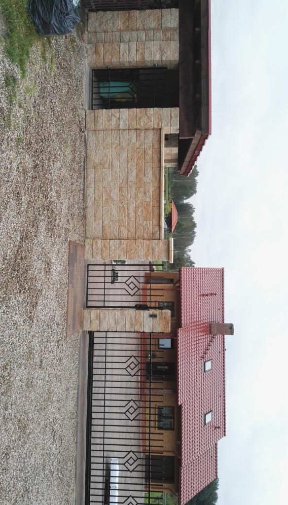 Ukończony dom jednorodzinny z czerwoną dachówką, ogrodzony czarnym, metalowym płotem z geometrycznym wzorem, elewacja wykończona płytkami imitującymi kamień.