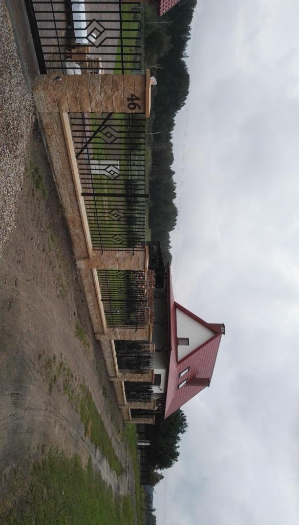 Nowoczesny dom z czerwonym dachem widziany zza ogrodzenia z kamiennymi słupkami z numerem 46, otoczony zielenią i pochmurnym niebem.