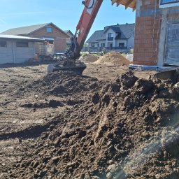 Firma-handlowo-usługow transportowa,,PE-TOM ''Krzysztof Pestka - Koparka nabiera ziemię na budowie domu jednorodzinnego. W tle sterta piasku i inne budynki. Widoczne ślady opon na ziemi.