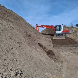Firma-handlowo-usługow transportowa,,PE-TOM ''Krzysztof Pestka - Czerwono-biała koparka O&K na tle sterty ziemi pod błękitnym niebem. Widoczne ślady opon i nierówności terenu. Sprzęt budowlany w trakcie prac ziemnych.