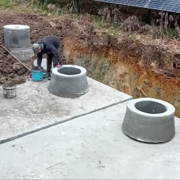 Firma-handlowo-usługow transportowa,,PE-TOM ''Krzysztof Pestka - Montaż betonowych kręgów studni na placu budowy. Widoczny wykop, częściowo ułożone płyty i osoba w trakcie pracy. W tle panele słoneczne.